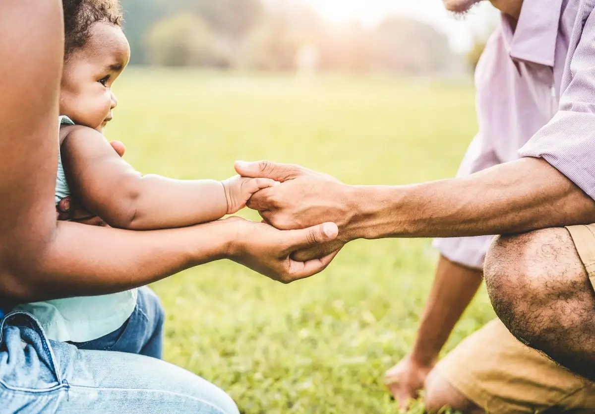 Close-up of parents holding their baby’s hands outdoors on a sunny day, symbolizing family support.